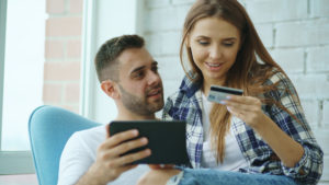 Young cheerful couple using digital tablet computer for online shopping sitting on balcony in modern loft apartment
